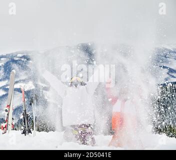 Zwei Skifahrer werfen frischen Pulverschnee hoch in die Luft. Mann und Frau haben Spaß im Skigebiet mit schönen Bergen im Hintergrund. Konzept der Wintersport-Aktivitäten, Spaß und Beziehungen. Stockfoto