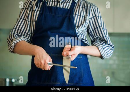 Der Koch steht in der Küche in einer blauen Schürze und einem gestreiften Hemd, hält ein Messer in der Hand und schärft es auf einem Wetzstein, um es schärfer zu machen Stockfoto