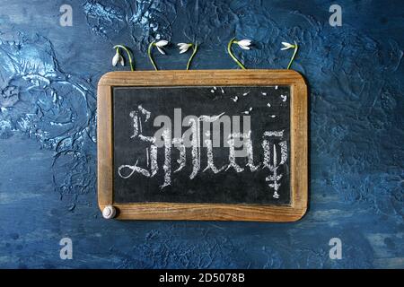 Kalligraphische Inschrift gothic Hand schrift Buchstaben Feder auf Schwarze Schiefertafel mit Blüte Schneeglöckchen über blaue Textur Hintergrund. Flach, Raum Stockfoto