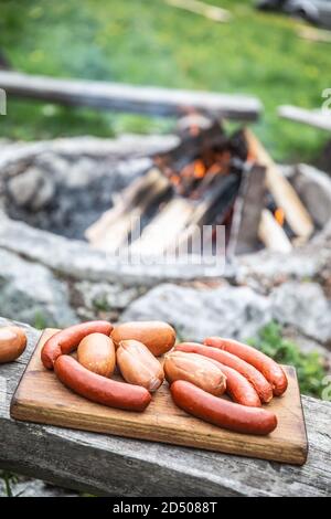 Würste auf einem hölzernen Schneidebrett bereit zum Braten Ein Lagerfeuer im Hintergrund Stockfoto