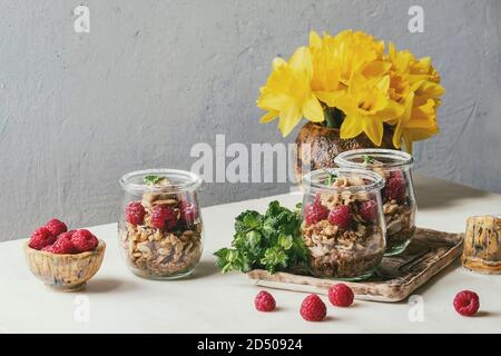 2-in-1-Dessert in Töpfen. Keks, Kaffeesahne, Muttern, Himbeeren, frischer Minze. Auf keramischen Fach mit Narzissen Blumen, Beeren und Grüns serviert über Stockfoto