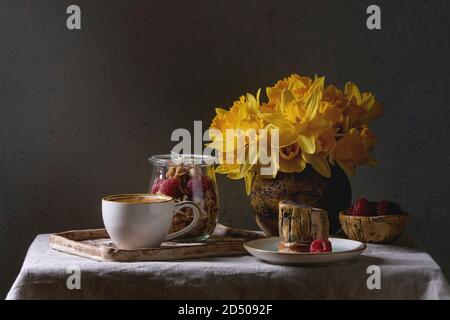 2-in-1-Dessert im Glas. Keks, Kaffeesahne, Muttern, Himbeeren, frischer Minze. Mit Tasse Kaffee Espresso, Beeren serviert, Narzissen Blumen und Kanne o Stockfoto