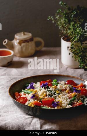 Couscous Salat mit parboiled Gemüse Karotten, grüne Bohnen, Mais, Spinat in Keramik Teller mit Tomaten, Sesam und essbare Blumen, Kaffee Stockfoto