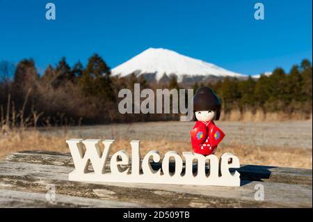 Schöne japanische traditionelle Kokeshi Puppe in roten Kimono gekleidet auf dem Wort "Willkommen". Hintergrund mit Fuji. Horizontale Aufnahme. Stockfoto