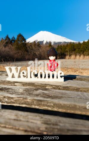 Schöne japanische traditionelle Kokeshi Puppe in roten Kimono gekleidet auf dem Wort "Willkommen". Hintergrund mit Fuji. Vertikale Aufnahme. Stockfoto