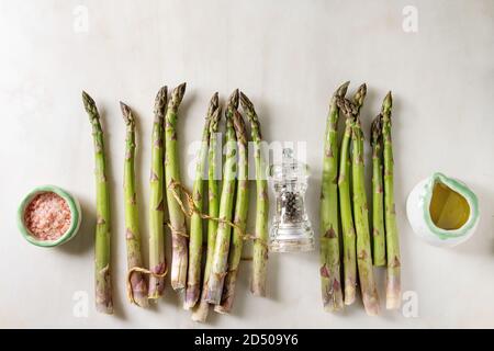 Bündel von organischen grüner Spargel mit Zutaten zum Kochen rosa Meersalz, schwarzer Pfeffer und Olivenöl in weißem Marmor Hintergrund. Flach, Raum Stockfoto