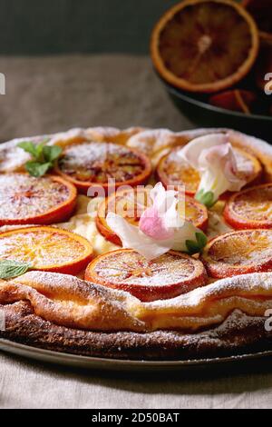 Hausgemachter Käsekuchen mit sizilianischer Blutorangen, dekoriert von essbaren Blüten, Minze und Zucker Pulver in der Platte mit geschnittenen Orangen oben g serviert. Stockfoto