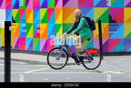 London, England, Großbritannien. Mann auf einem Santander Hire Cycle (Boris Bike) in Long Acre, vorbei an einem bunten Bauplatz. Stockfoto