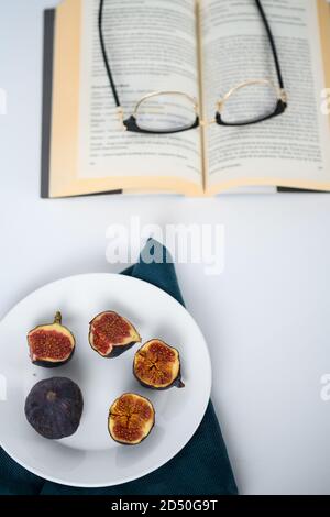 Feigen auf einem weißen Teller, ein Paar Gläser auf einem offenen Buch Stockfoto