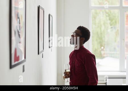 Seitenansicht Porträt eines jungen afroamerikanischen Mannes, der Gemälde ansieht und Sektglas bei der Eröffnung der Kunstgalerie hält, Kopierraum Stockfoto