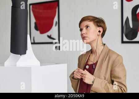 Porträt einer zeitgenössischen jungen Frau, die Skulpturen ansieht und Audioguide in der Kunstgalerie, Copy Space, hört Stockfoto