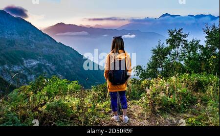 Junge blonde Frau von hinten in gelbem Hoodie mit Rucksack blickt auf einen hellen Sonnenuntergang in den Bergen. Trekking in den Bergen, aktive Lebensweise, h Stockfoto