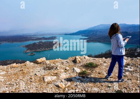 Junge Frau mit im Wind flatternden Haaren fotografiert den wunderschönen blauen Slansko See in Montenegro. Landschaft. Mädchen von hinten Stockfoto