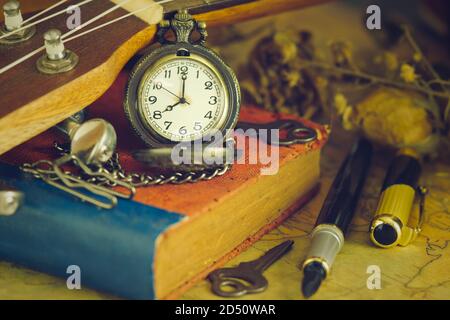 Eine antike Taschenuhr lehnte sich an eine Ukulele und ein altes Buch mit Vintage-Karte und Messingstift auf Holztisch platziert. Nahaufnahme und Kopie Platz für Text. Stockfoto