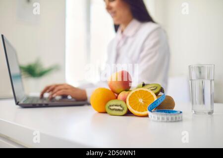 Obst, Maßband und Glas Wasser auf dem Schreibtisch gegen verschwommene Ernährungsberater arbeiten auf Laptop platziert Stockfoto