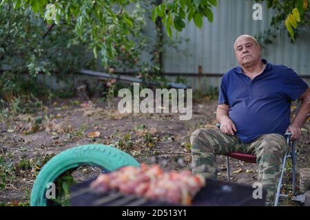 Nahaufnahme der Spieße mit rohem Schaschlik und Gemüse auf Holzgrill. Reifer Mann rösten saftigen Grill im Garten. Konzept von Picknick im Freien. Stockfoto