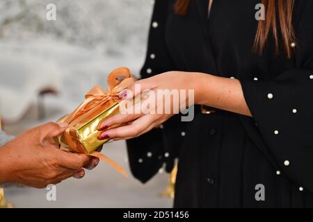Nahaufnahme der weiblichen Hände, die eine goldene Geschenkbox für eine andere Frau auf einem unfokussieren Hintergrund geben. Weihnachts- und Geschenkkonzept. Stockfoto