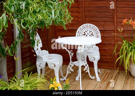 Attraktiver weißer Bistrotisch und Stühle draußen auf Holz Terrasse, umgeben von Topfpflanzen und Weidenbäumen mit Orange Und gelbe Tigerlilie Stockfoto