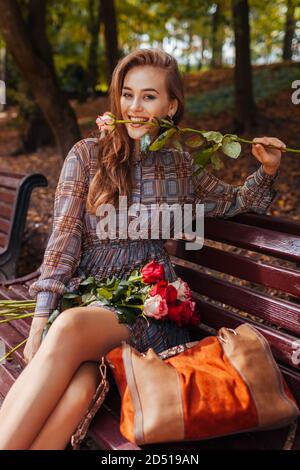 Schöne Frau sitzt auf der Bank mit Strauß Rosen im Herbst Park. Mädchen mit Spaß beißen Rose Stockfoto