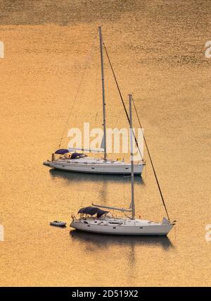 Segelboote bei Sonnenuntergang, Sonnenaufgang. Zwei weiße Yachten liegen mitten im goldenen, ruhigen Meer. Kopierbereich, vertikales Foto. Stockfoto