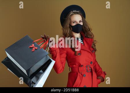 Hallo Herbst. Glückliche moderne 40 Jahre alte Frau in rotem Mantel mit schwarzer Maske und Papier Einkaufstaschen isoliert auf braunem Hintergrund. Stockfoto