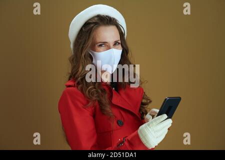 Leben während einer Coronavirus-Pandemie. Porträt der trendigen Frau in rotem Mantel Senden SMS-Nachricht mit Smartphone auf beigem Hintergrund. Stockfoto