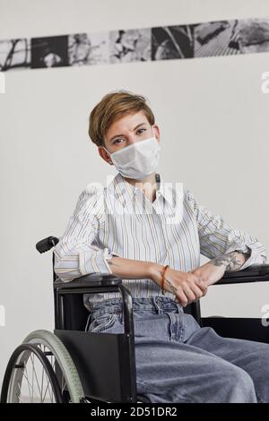 Vertikales Porträt einer jungen Frau mit Rollstuhl und Maske Beim Blick auf die Kamera gegen die weiße Wand in der modernen Kunst Galerie Stockfoto