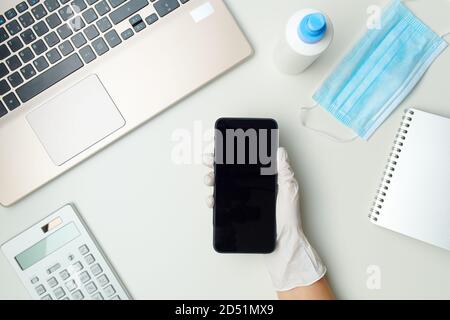 Obere Ansicht der weiblichen Hände in weißen Gummihandschuhen zeigt Smartphone leeren Bildschirm in der Nähe von medizinischen Maske, Desinfektionsmittel und Laptop auf weißem Tisch. Stockfoto