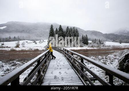 Frau in gelber Jacke auf der Brücke im Naturschutzgebiet Zelenci Wenn der erste Schnee der Saison fällt, Slowenien Stockfoto