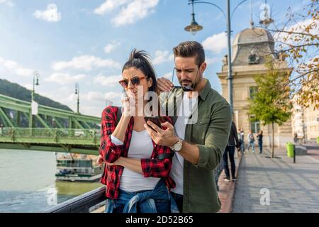 Ein junges Paar streitet über den Inhalt, der in einer europäischen Stadt auf dem Handy-Bildschirm zu sehen ist. Stockfoto