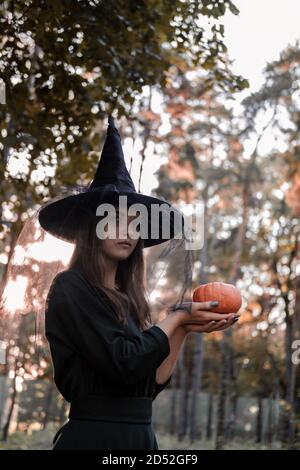 Junge Frau in dunklem Kleid und Hexenhut hält einen Kürbis in den Händen. Halloween Party Kostüm. Wald, Park mit Herbstbäumen Stockfoto