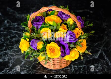 Schöner Strauß Rosen auf schwarzem Marmor. Gelbe und blaue Blüten in einem Weidenkorb. Ungewöhnliche Zusammensetzung der gesammelten violetten und gelben Blüten. Stockfoto