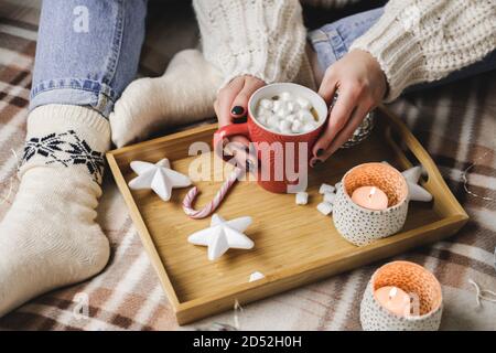 Die junge Frau sitzt auf Karos im kuscheligen Strickpullover aus Wolle und hält eine Tasse Kakao mit Marshmallows. Hygge Neujahr, gemütliche Weihnachten, Vorbereitung auf den Urlaub Stockfoto