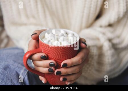 Die junge Frau sitzt auf Karos in kuschelig gestricktem weißen Wollpullover und hält eine Tasse Kakao mit Marshmallows in den Händen. Hygge Neujahr, gemütliche Weihnachten, Stockfoto