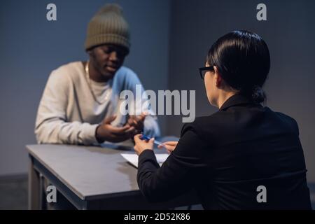 Ein Anwältin mit Brille und ein junger schwarzer Kerl kommunizieren im Verhörraum über die Gründe für seine Inhaftierung. Stockfoto