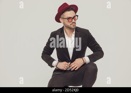 Selbstbewusstes Modelmodell, das seine Jacke anpasst, trägt Sonnenbrille und Hut, während es auf grauem Studiohintergrund hocket Stockfoto