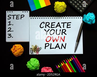Weiße Notizen mit der Aufschrift 'Create your own Plan' und 'Step 1, 2, 3' auf schönem schwarzen Tisch, farbigem Papier, Buntstiften, Büroklammern, Stift und Stockfoto