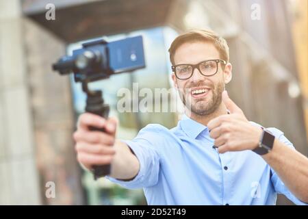Junge fröhliche männliche Blogger trägt blaues Hemd und Brillen mit einem Gimbal mit Smartphone, lächelnd und zeigt Daumen nach oben, während die Aufnahme neuer Video Stockfoto