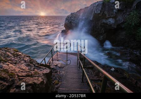 Treppen führen hinunter zum Meer mit großen Wellen und Sonnenuntergang im Hintergrund Stockfoto