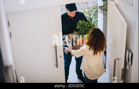 Blick aus der Perspektive einer Kundin, die Lebensmittel vom Lieferer vor der Haustüre erhält. Liefermitarbeiter liefert eine Schachtel Obst und Gemüse Stockfoto