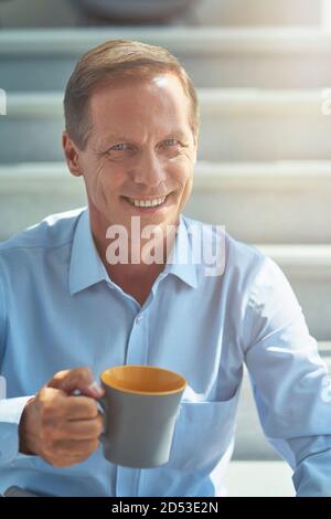 Glücklicher reifer Geschäftsmann im Hemd hält Tasse Kaffee, Blick auf die Kamera und lächeln, während eine Pause im Büro, vertikale Aufnahme Stockfoto
