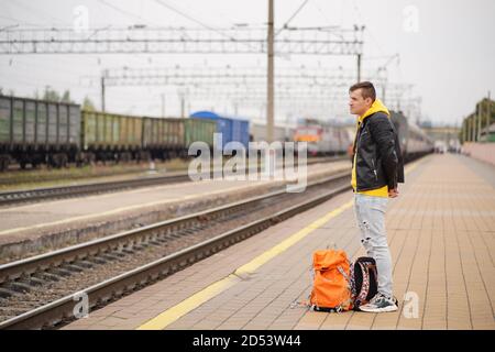 Der junge Mann steht auf dem Bahnsteig und wartet auf den Zug. Männlicher Passagier mit Rucksäcken auf Bahnsteig in Warten auf Zugfahrt. Konzept des Tourismus Stockfoto