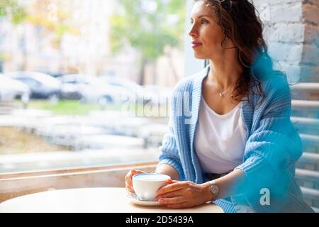 Unfokussierte glückliche Frau mit langen welligen Haaren in warmen Pullover Tasse heißen blauen Latte halten Stockfoto