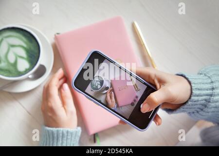 Frau fotografiert blauen Kaffee Latte, rosa farbigen Planer 2021 und Gold-Stift auf ihrem Smartphone. Stockfoto