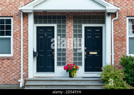 Zwei schwarze Metalltüren mit einem roten Blumentopf zwischen den beiden in der Mitte eines Gebäudes. Es hat eine rote Ziegelwand mit weißen Holzverkleidung. Stockfoto