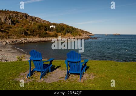 Zwei blaue Adirondack-Stühle umgeben von Gras mit Blick auf das Meer und einen kleinen Strand. Die Stühle sind leer. Stockfoto