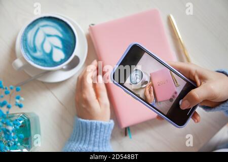 Frau fotografiert blauen Kaffee Latte, rosa farbigen Planer 2021 und Gold-Stift auf ihrem Smartphone. Stockfoto