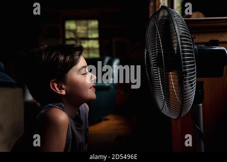 Kleiner Junge, der sich vor einem elektrischen Ventilator im dunklen Zimmer abkühlte. Stockfoto