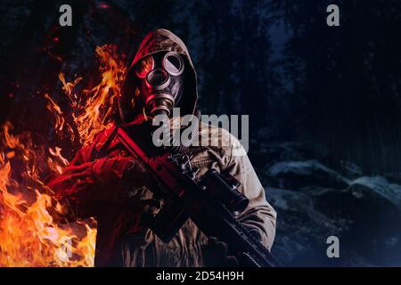 Foto eines postapokalyptischen Stalker Soldaten in Gasmaske und Kapuzenjacke halten Gewehr und stehend und brennend auf dunklen Nachtwald Hintergrund. Stockfoto