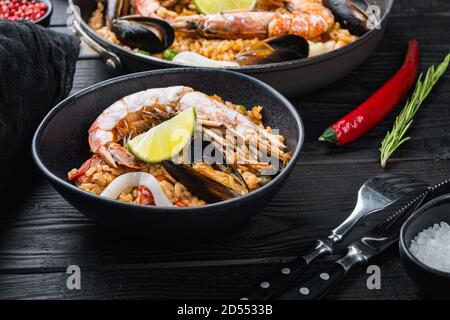 Hausgemachte spanische Paella mit Meeresfrüchten in Bratpfanne und schwarzer Schüssel Auf schwarzem Holzhintergrund Stockfoto